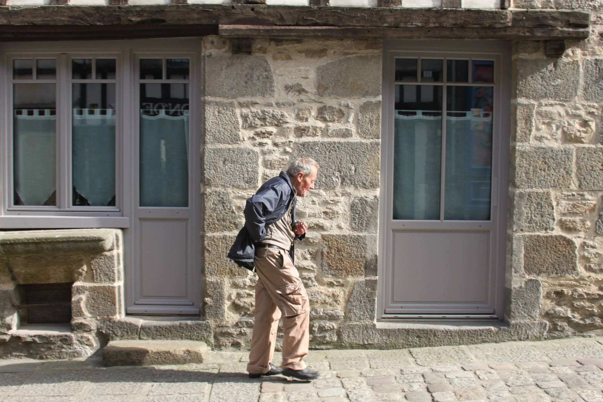 A man in Dinan on his evening walk. He is passing between two windows separated by a stone wall.