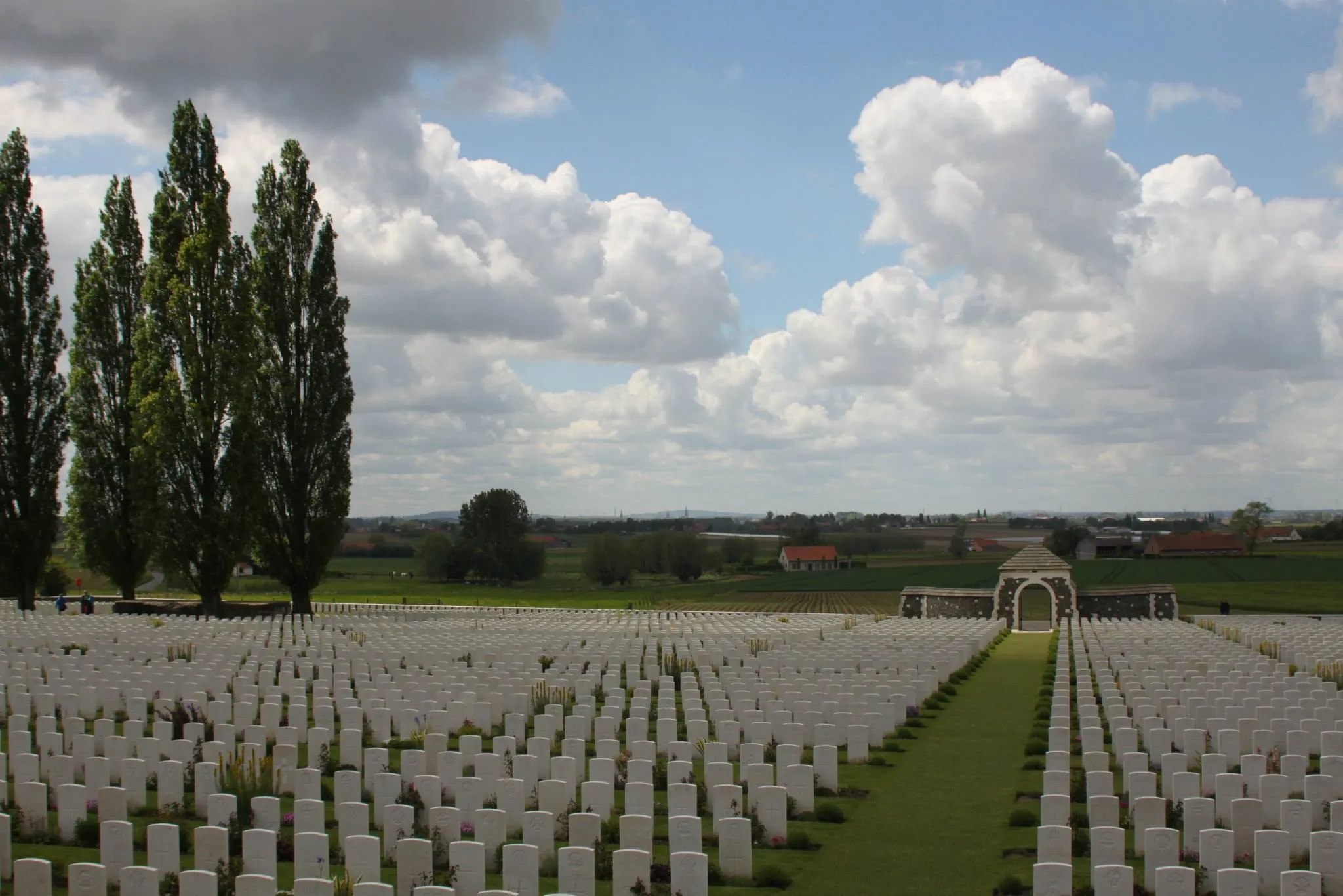 Tyne Cot Cemetery looking down the aisle over hundreds of headstones facing towards the main entrance, beyond which lies the rolling hills of Flanders dotted with farmhouses.