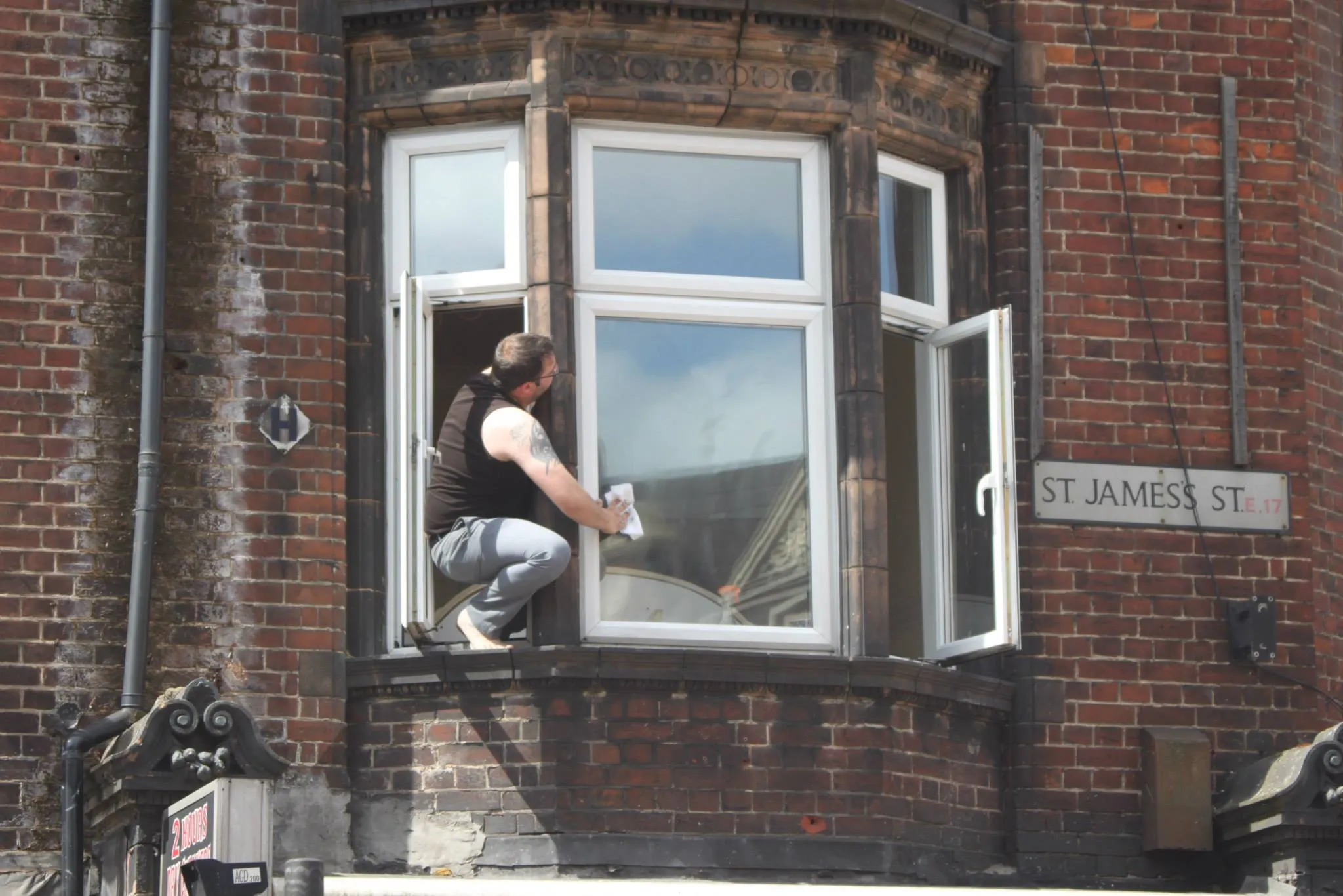 A man washes his window on St. James's St. leaning precariously out of an open window to do so.